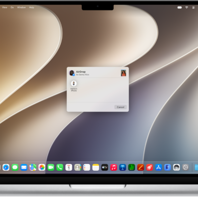 A Mac is next to an iPhone. The AirDrop dialog is open on the Mac desktop, ready to share a document with the iPhone. The iPhone screen has a notification about receiving the document.