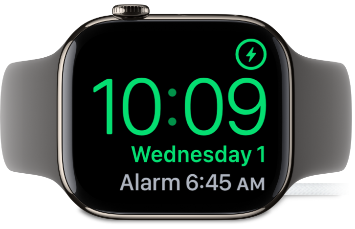 An Apple Watch placed on its side and connected to the charger, with the screen showing the charging symbol in the top-right corner, the current time below that, and the time of the next alarm.